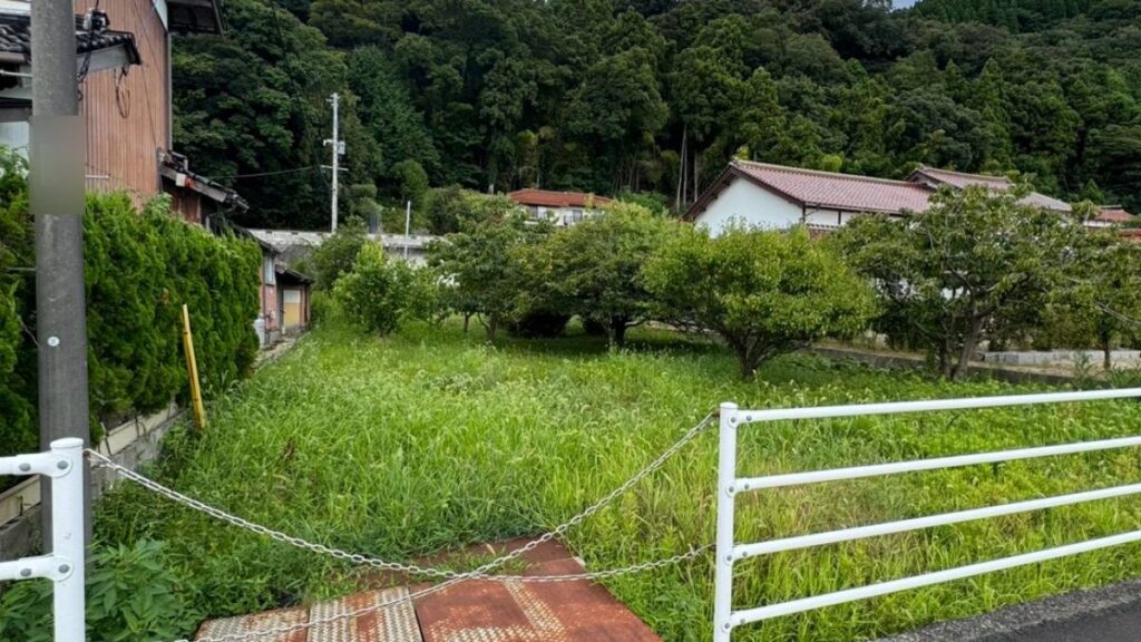 白いガードパイプで囲われた草だらけの空き地と背後の集落